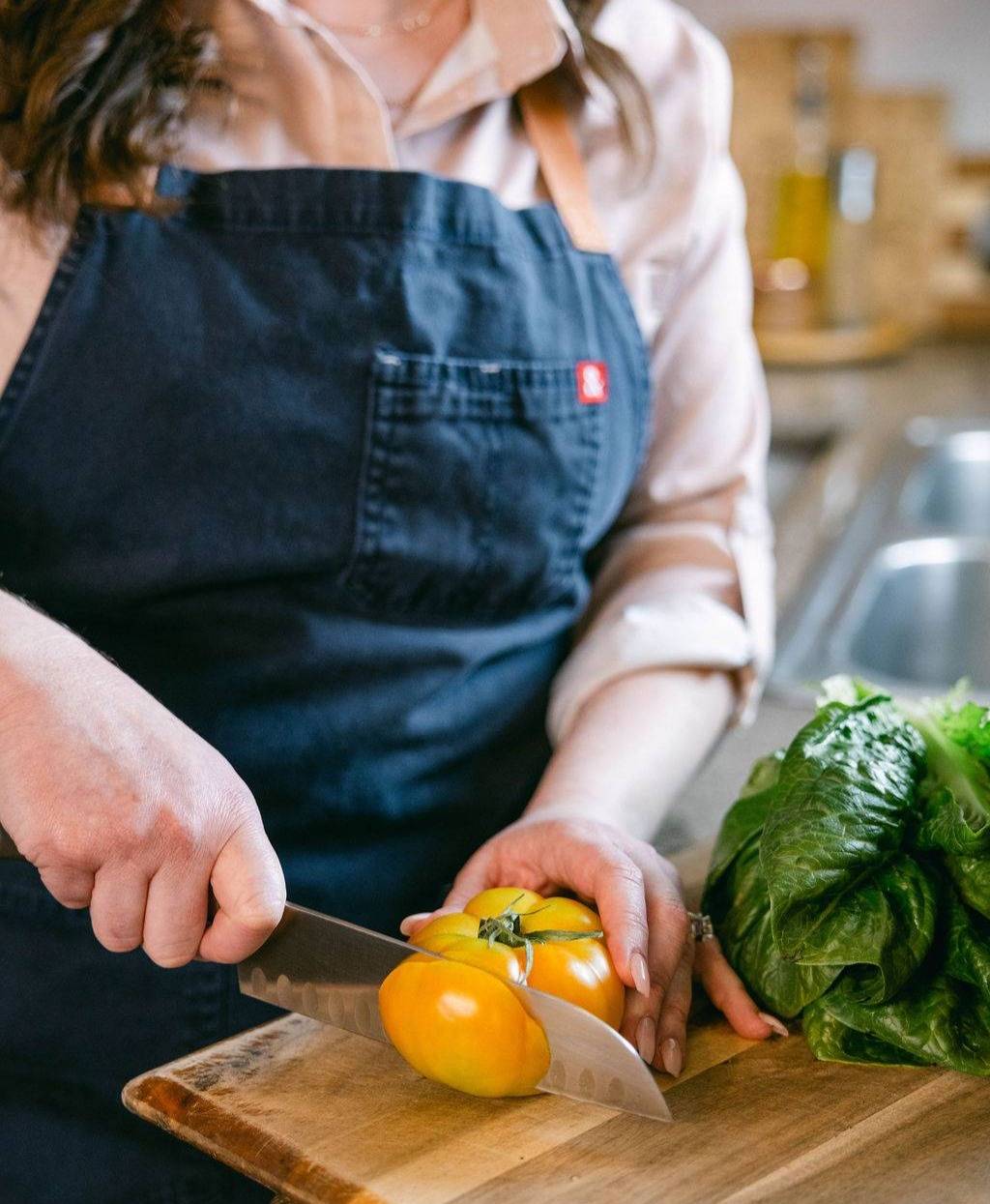 A person wearing a dark apron is slicing a large yellow tomato on a wooden cutting board. Next to the tomato, there is a bunch of fresh green lettuce. The scene is set in a kitchen with a slight view of a sink and counter in the background.