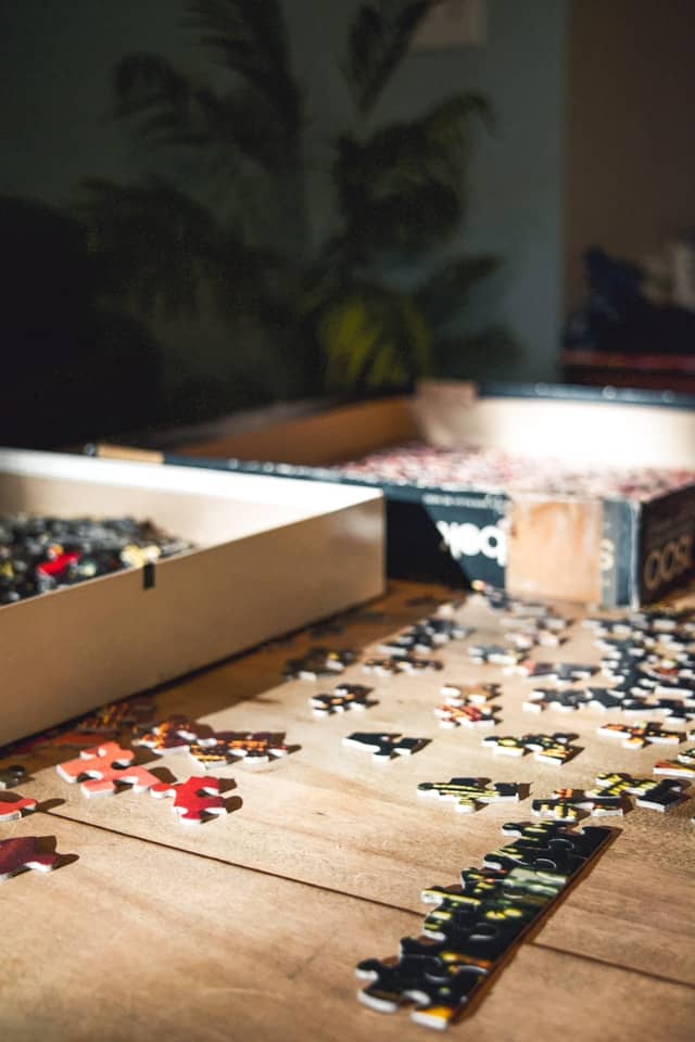 A cozy domestic scene shows multiple jigsaw puzzles in various stages of completion spread across a light wooden table. In the foreground, loose puzzle pieces are scattered on the tabletop, while partially completed sections reveal what appears to be a floral or nature-themed image. Two puzzle boxes rest nearby—one with its lid open showing remaining pieces inside. The shallow depth of field creates a soft bokeh effect in the background, where a leafy plant pattern on what might be wallpaper or fabric becomes an artistic blur of dark greens and yellows.