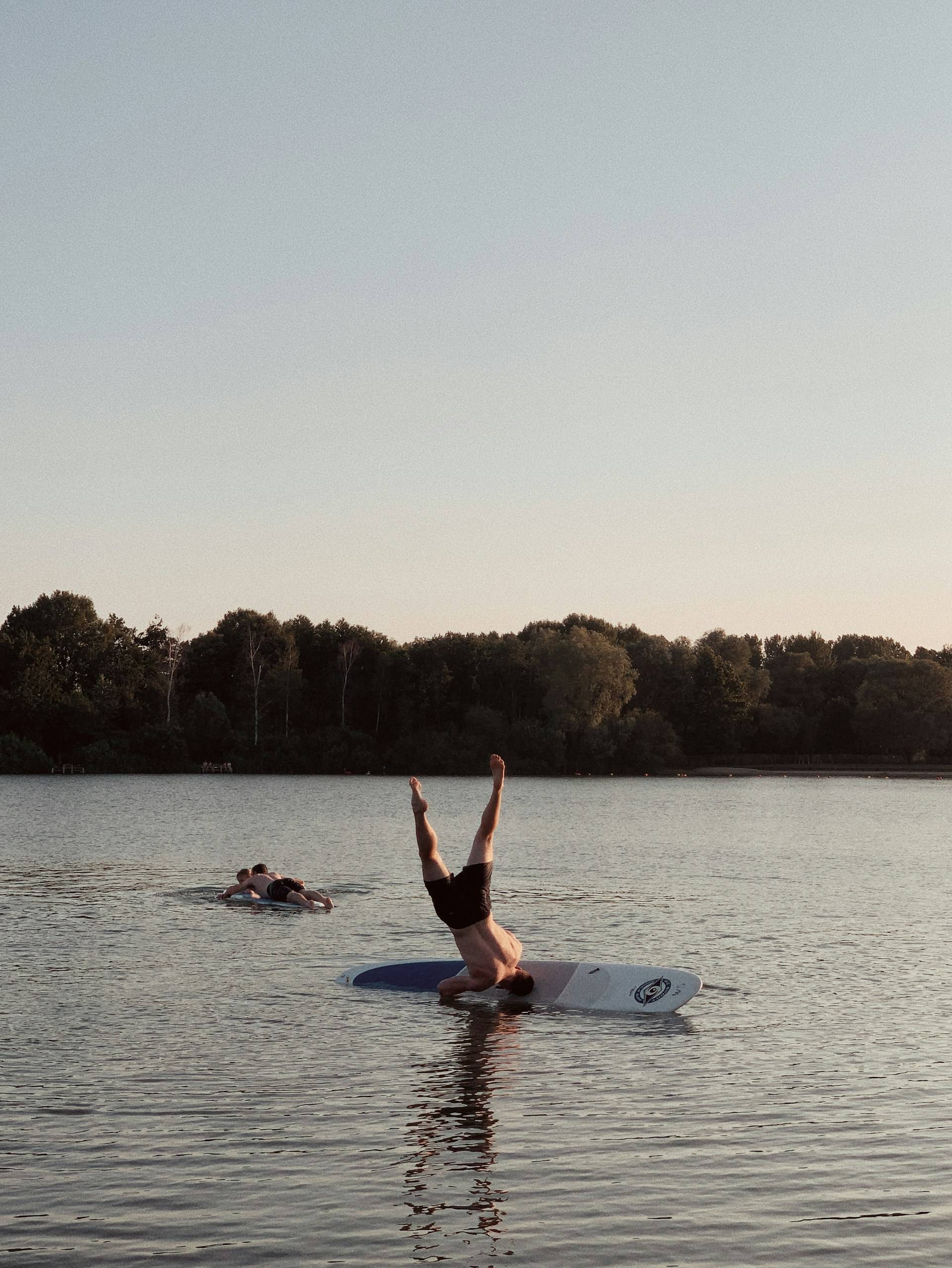 A person attempts to do a headstand while on white paddleboard in the middle of a tranquil lake, their legs pointing skyward in defiant celebration of summer's physics-bending possibilities. However, the headstand is unsuccessful and they are falling into the water. The calm water mirrors their inverted silhouette, creating ripples that stretch toward the dense wall of trees standing sentinel along the distant shore. 