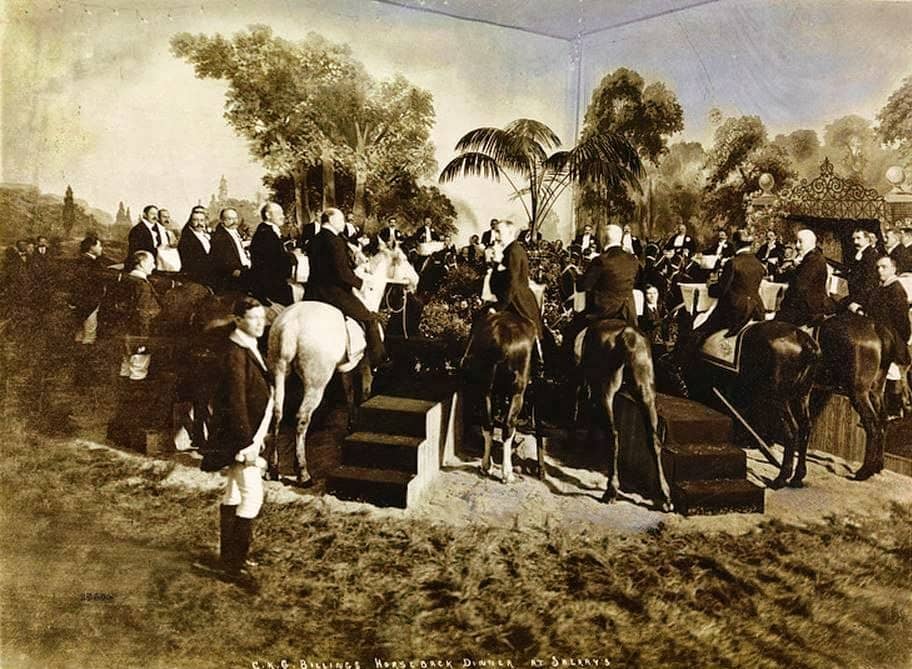 Tinted vintage photograph from 1903 of C.K.G. Billing's infamous dinner party. A group of male guest in white tie attire sit atop horses for the extravagent feast. Each diner/horse was assigned a groom to attend to them during the meal. 