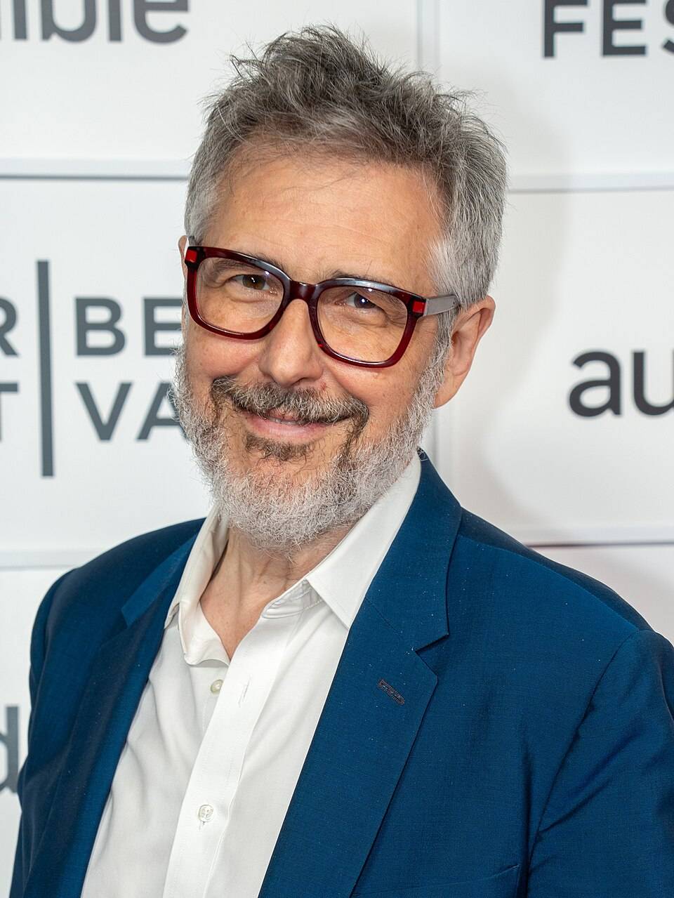 Photograph of Ira Glass, a 66 year-old man, at the Tribeca Film Festival. He wears a crisp white shirt and dark blue blazer. His signature eyeglasses are in a dark brown color. His hair and beard are shades of white, silver, and grey. 