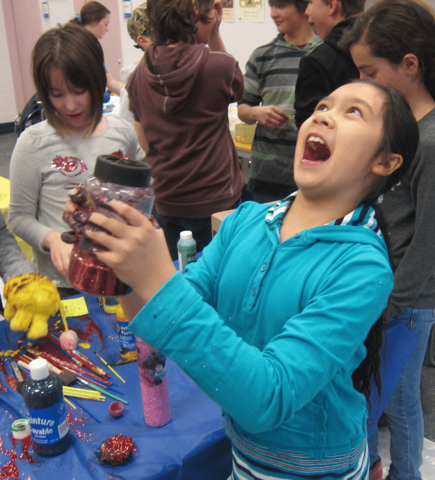 A crowded indoor event space at a public library in Ontario. A kid who looks to be about 10 years old is holding a giant shaker of glitter in paint and glue-covered hands and is maniacally laughing. 