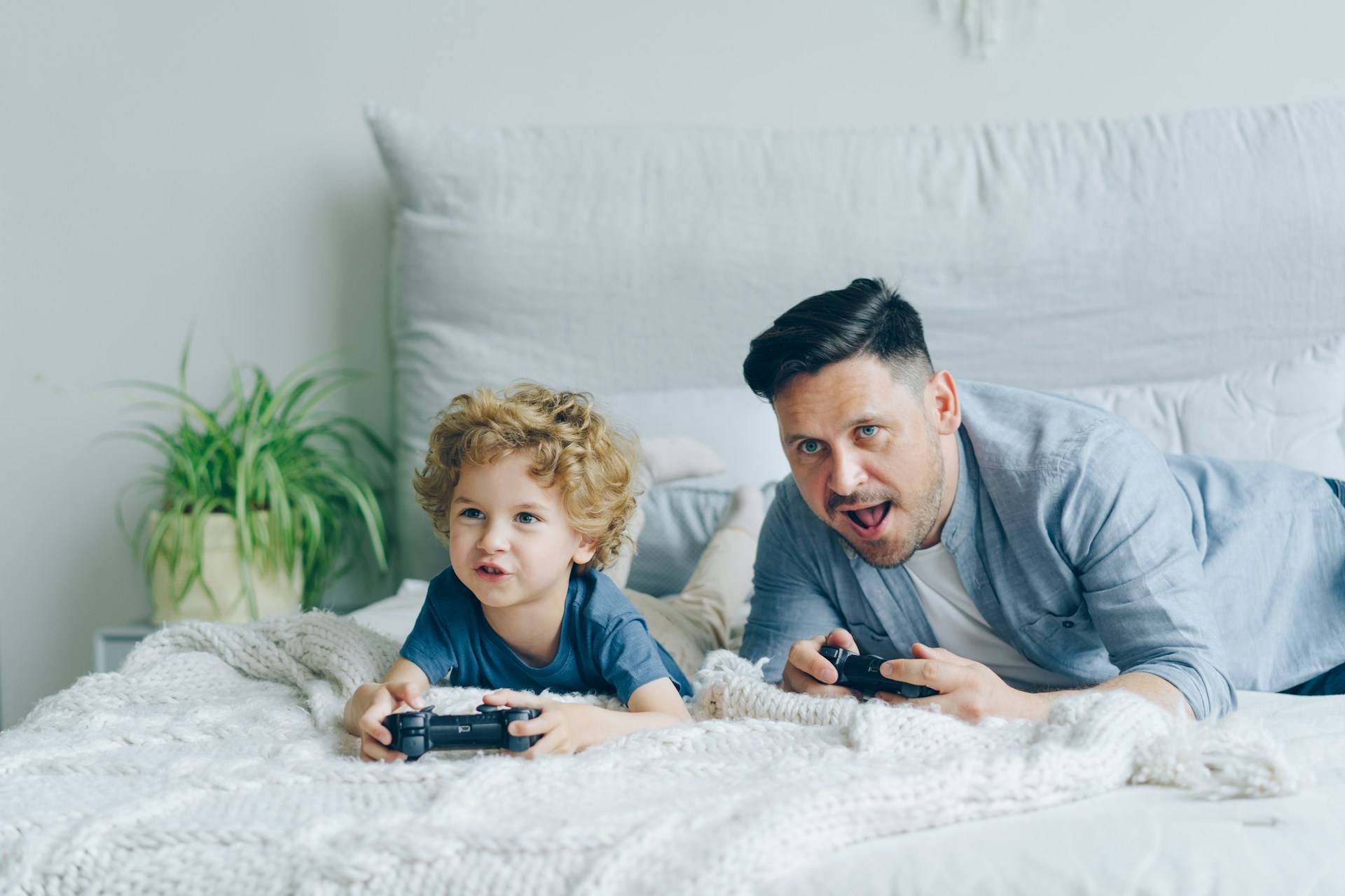A father and a little boy lay across a white bedspread with video game controllers in the their hands. It's clear from the expressions on their faces that the father is animatedly engaging with the delighted child. 