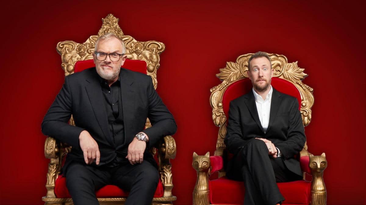 Official publicity still from Channel 4 for Taskmaster. Greg Davies, a large man with glasses dressed all in black sits on a throne. A slightly smaller chair is provided for Alex Horne, making a slightly terrified face next to Greg's authority. 