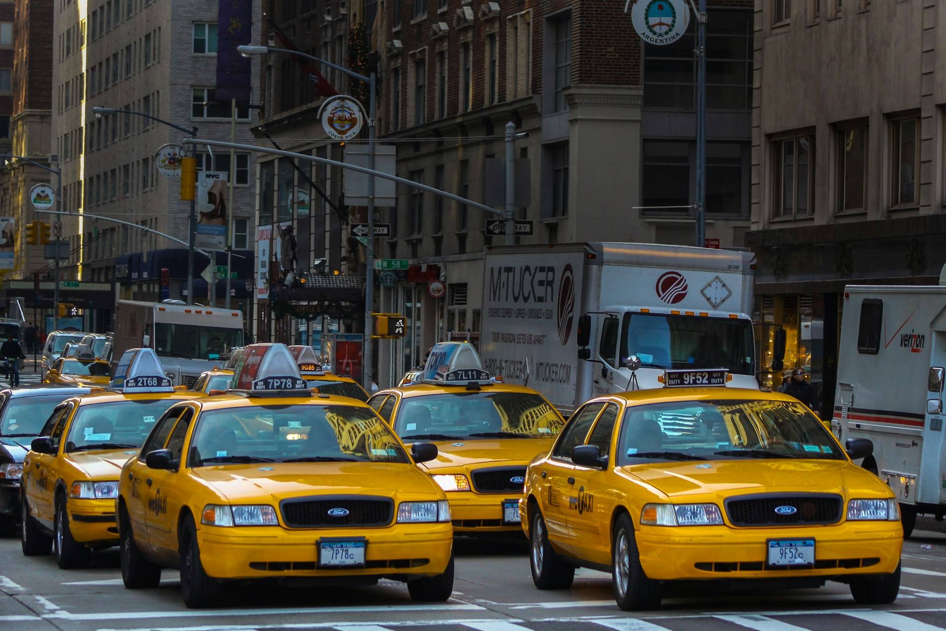 A quintessential New York City traffic scene with three iconic yellow taxi cabs lined up bumper-to-bumper on a busy street, surrounded by delivery trucks and urban architecture. The taxis display their distinctive medallion numbers on their roofs and Ford emblems on their grilles. Behind them, city buildings rise up with shop awnings and street signs visible in the urban canyon.