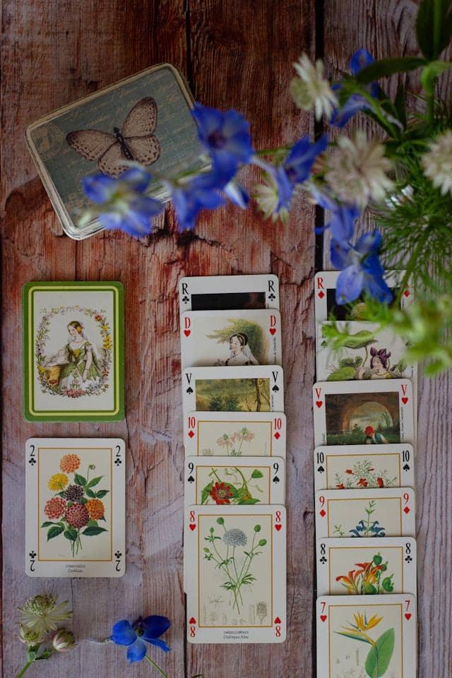 A dreamy arrangement of vintage botanical playing cards fans across weathered wooden planks, creating an artistic collision of nature and nostalgia. The cards feature delicate illustrations—a woman in a garden surrounded by flowers, vibrant dahlias and zi