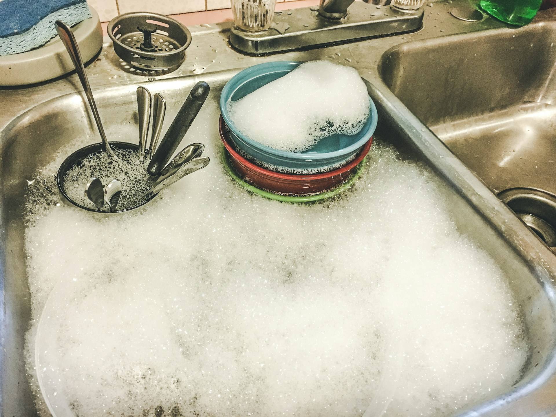 Photo of a kitchen sink with two metal basins. The left basin is full of soapy water and some colorful bowls and a cup of silverware is soaking in the water. 