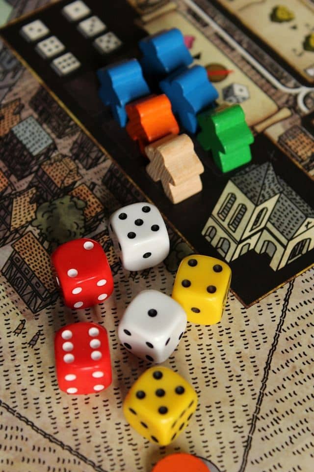 A vibrant tabletop gaming scene captures the essence of analog play. Colorful wooden meeples—cylindrical game pieces in bright blue, orange, green, and natural wood—are clustered on an illustrated game board featuring intricate drawings of medieval buildings and settlements. In the foreground, six dice are scattered across the board: two red dice, two white dice, and two yellow dice, each showing different numbers of dots. The composition celebrates the tactile joy of board gaming, where strategy meets chance in a riot of primary colors against vintage-style game art.
