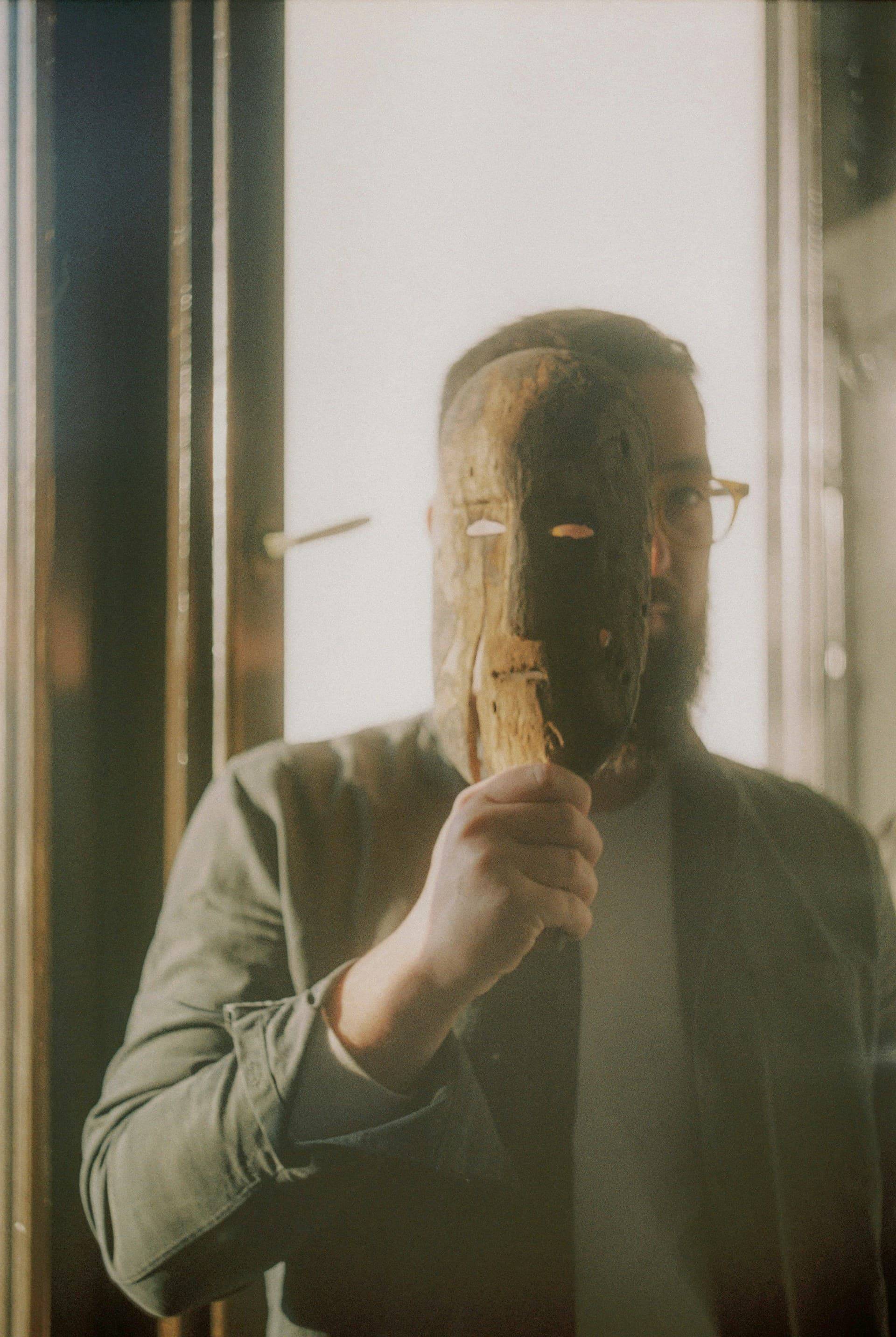  holds an ornate wooden mask with carved features up to their face, photographed in warm, natural lighting near a window. The mask appears to have traditional or theatrical styling with defined facial features.