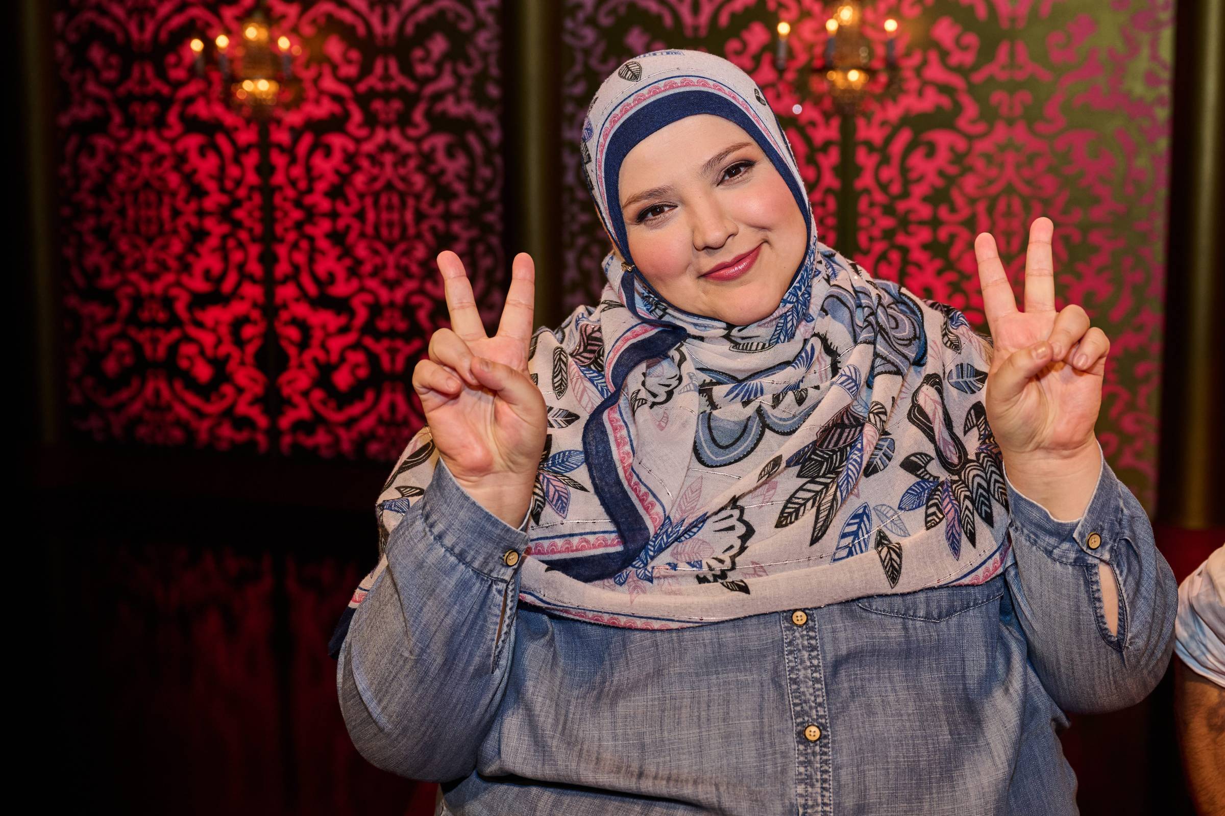 Photograph of Fatiha El-Ghorri in the Taskmaster Studio. A plus-size young Moroccan woman wears a denim shirt and a floral printed hijab. She has rosy pink cheeks and pink lipstick. 