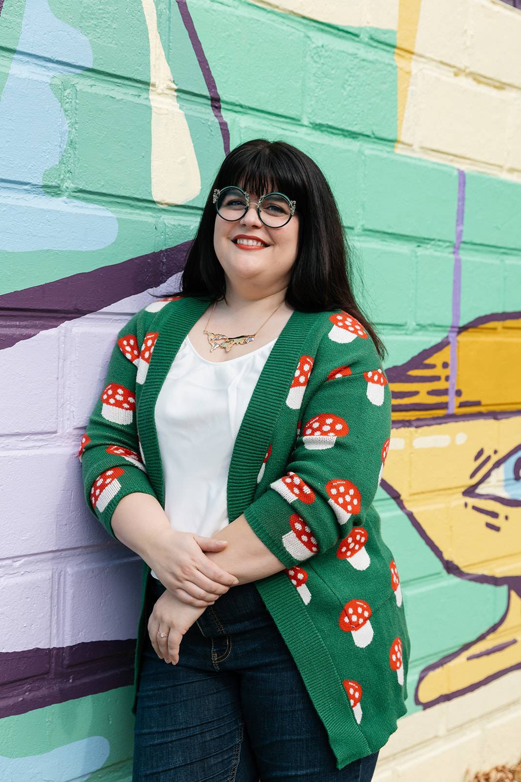 Photograph of Liz Gotauco, a middle-aged woman with dark hair and heavy bangs, leaning against a colorful wall mural. Liz is wearing round, green eyeglasses and a green cardigan sweater featuring cute toadstools. 