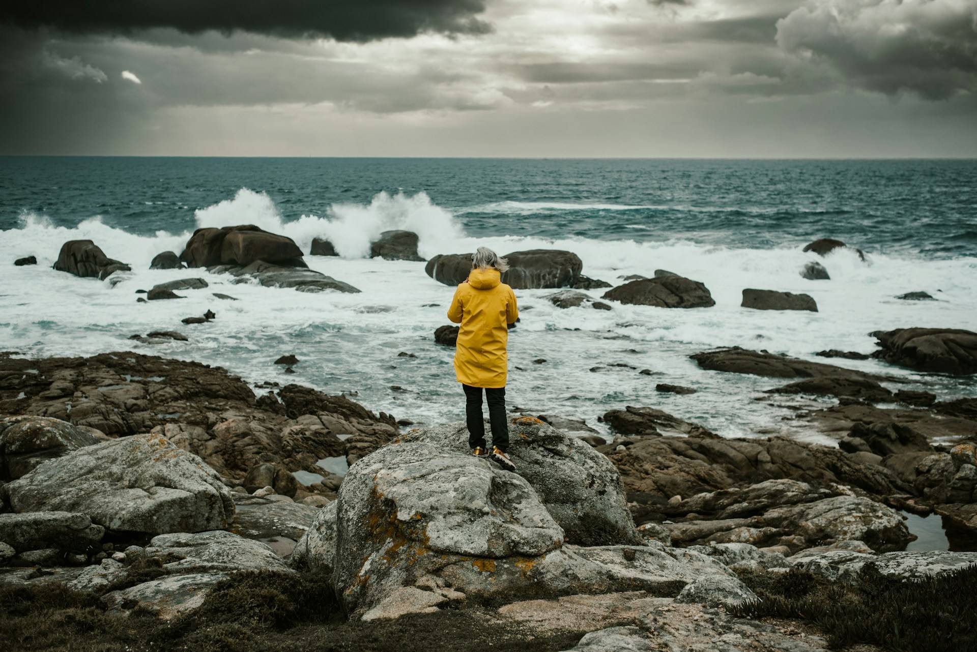 A solitary figure in a bright yellow raincoat stands defiantly on a rocky shoreline, facing a turbulent teal-gray sea. Dramatic storm clouds hover overhead, casting an ethereal light across the water. Waves crash against dark boulders, sending up sprays of white foam. Photo credit: Maksym Kaharlytskyi/Unsplash.