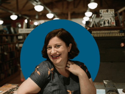 Photo composite of Nancy Martira, middle aged white woman with dark, red brown hair standing in a bookstore in Seattle. The overhead lights and shelves of books are visible behind her. She appears in front of a bold blue accent color. 
