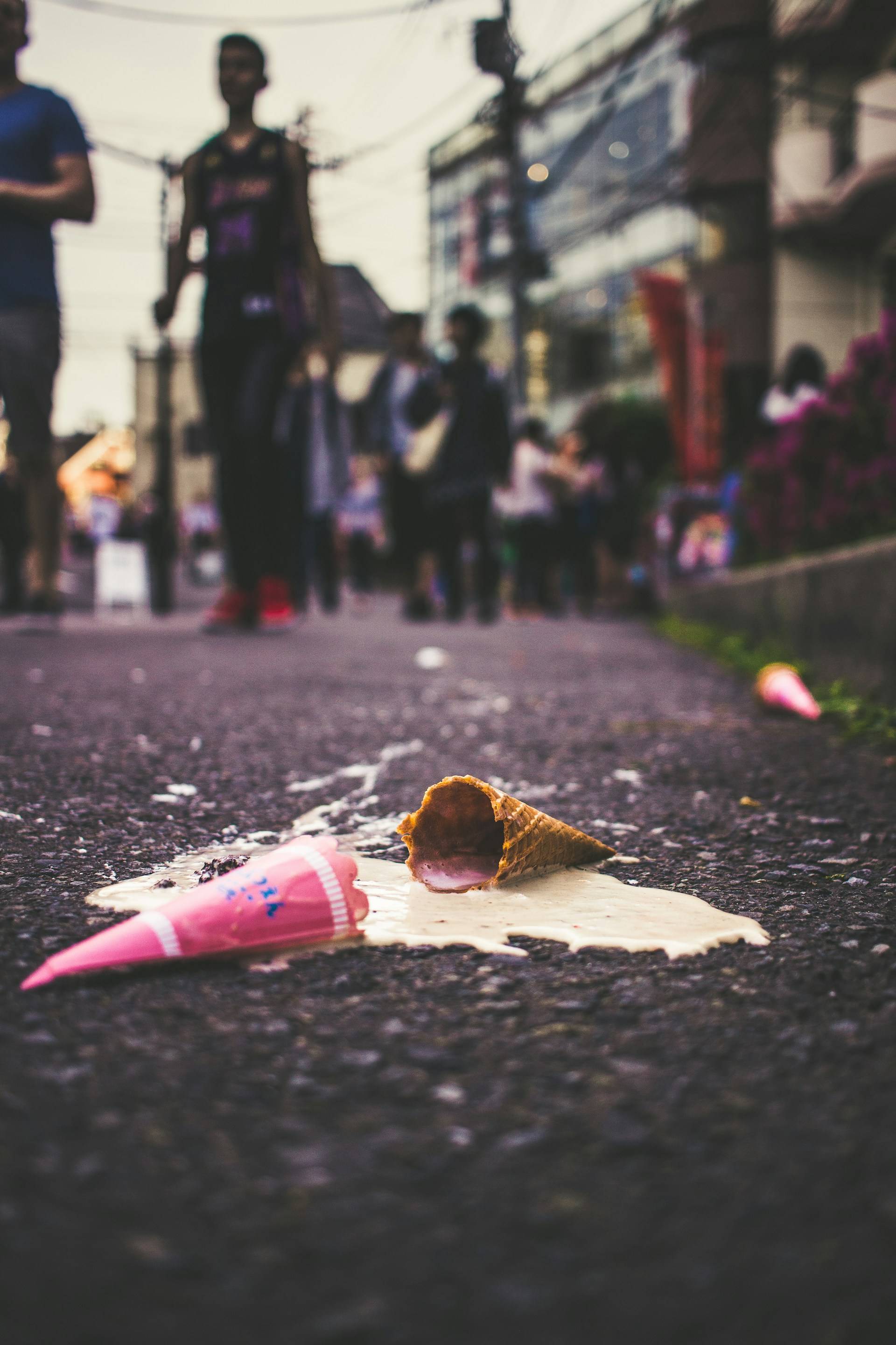 A dropped ice cream cone lies defeated on dark asphalt, its pink contents spilling dramatically onto the pavement. The cone rests on its side, a small pool of melting dessert forming beneath it, while the bright pink scoop with blue sprinkles has separated from its waffle home. In the blurred background, pedestrians continue their urban journey, oblivious to this small tragedy unfolding at street level.