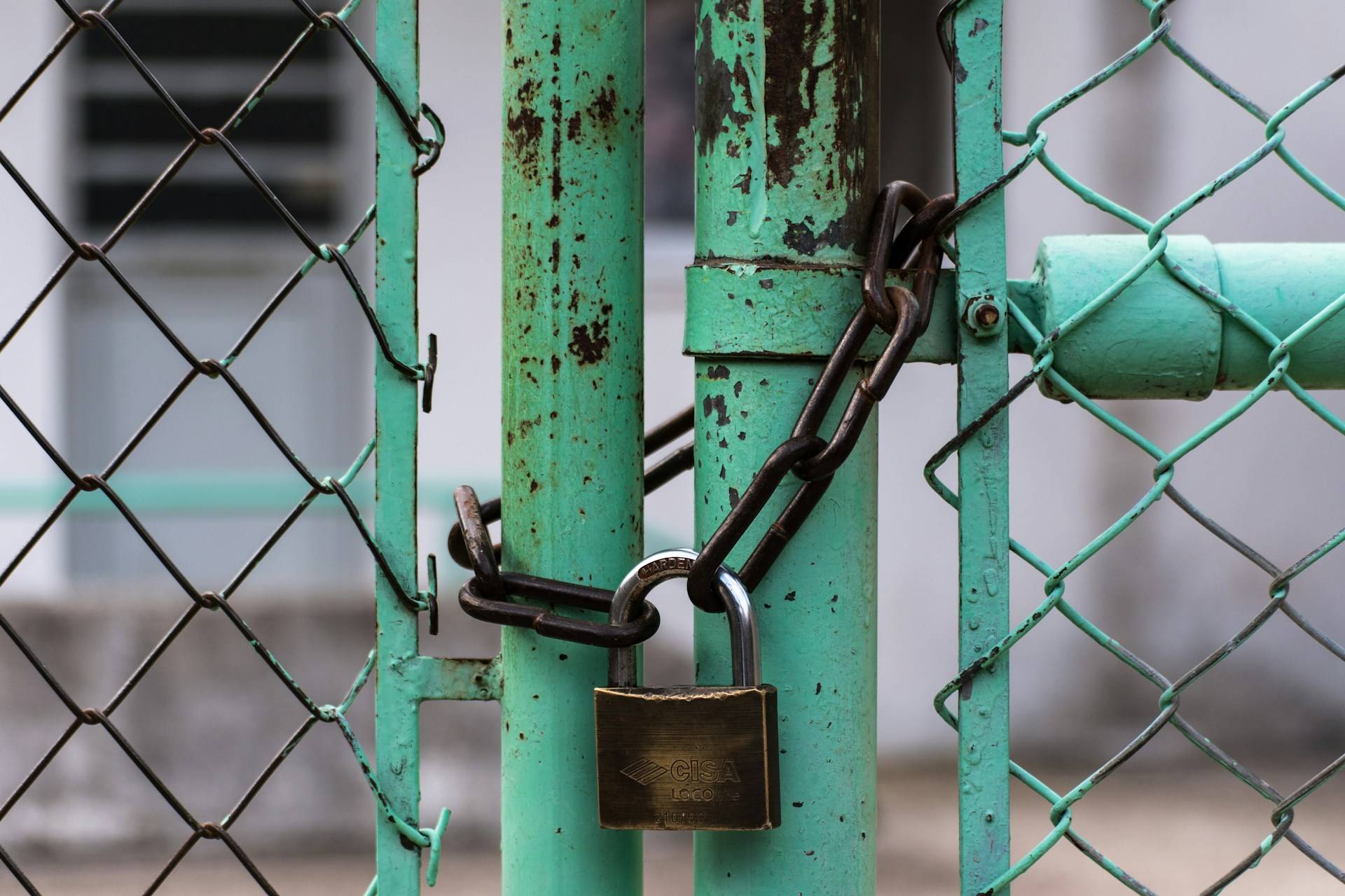A close-up photograph of a chain-link fence gate secured with a brass padlock and metal chain. The gate frame and posts are painted mint green, showing significant weathering with rust spots and peeling paint visible throughout the surface. The padlock is
