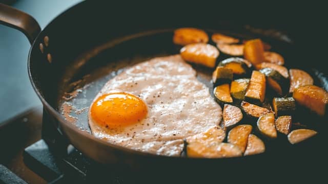 A close-up of a cast iron skillet on a stovetop containing: a sunny-side-up egg with its vibrant yellow yolk glistening on the left side, and golden-brown cubes of roasted sweet potato and squash clustered on the right. The egg appears to slide effortlessly on the pan's surface while the caramelized vegetables stick to their side – a perfect visual metaphor for Hanson's quote about how our brains process different experiences. Photo credit: Rob Wicks/Unsplash