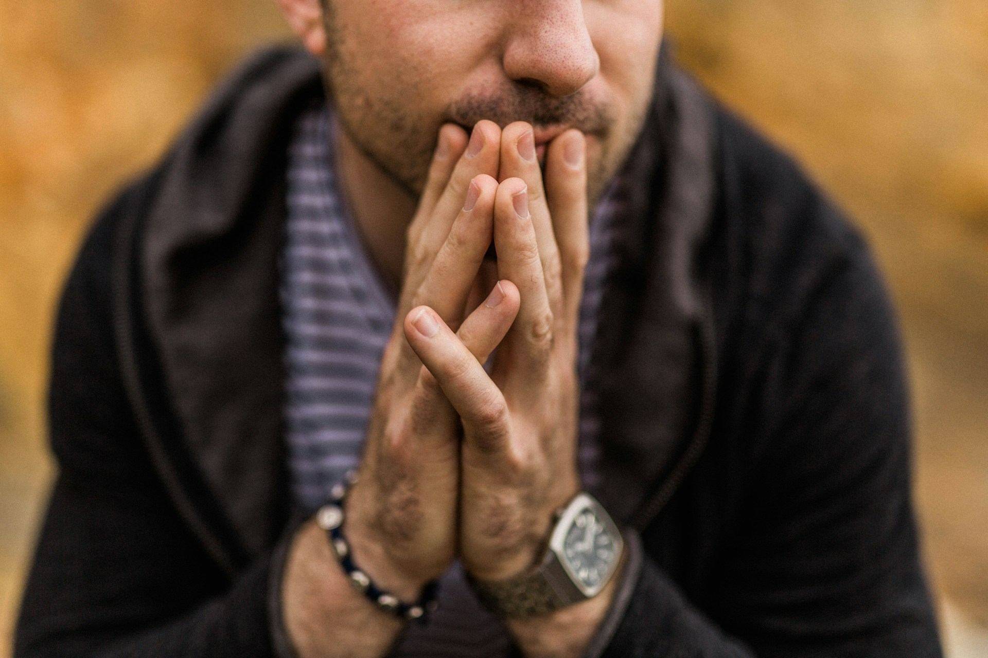 A man with brown skin and a light beard and mustache sits in quiet contemplation. We do not see his eyes, only the lower half of his face and his hands brought up to his chin. He wears a hooded sweatshirt over a striped shirt. One one wrist is a hemp bracelet and on the other wrist is a watch. 