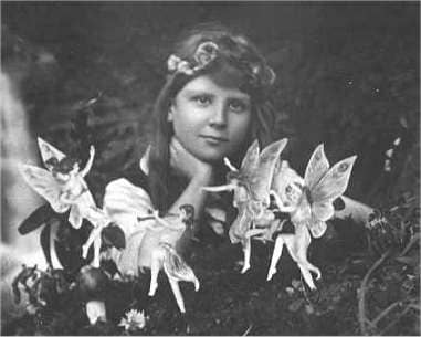 Black and white photograph taken in 1917. A young girl wears a homemade flower crown and gazes into the distance in a garden. In the foreground, "faires" frolic around her. 