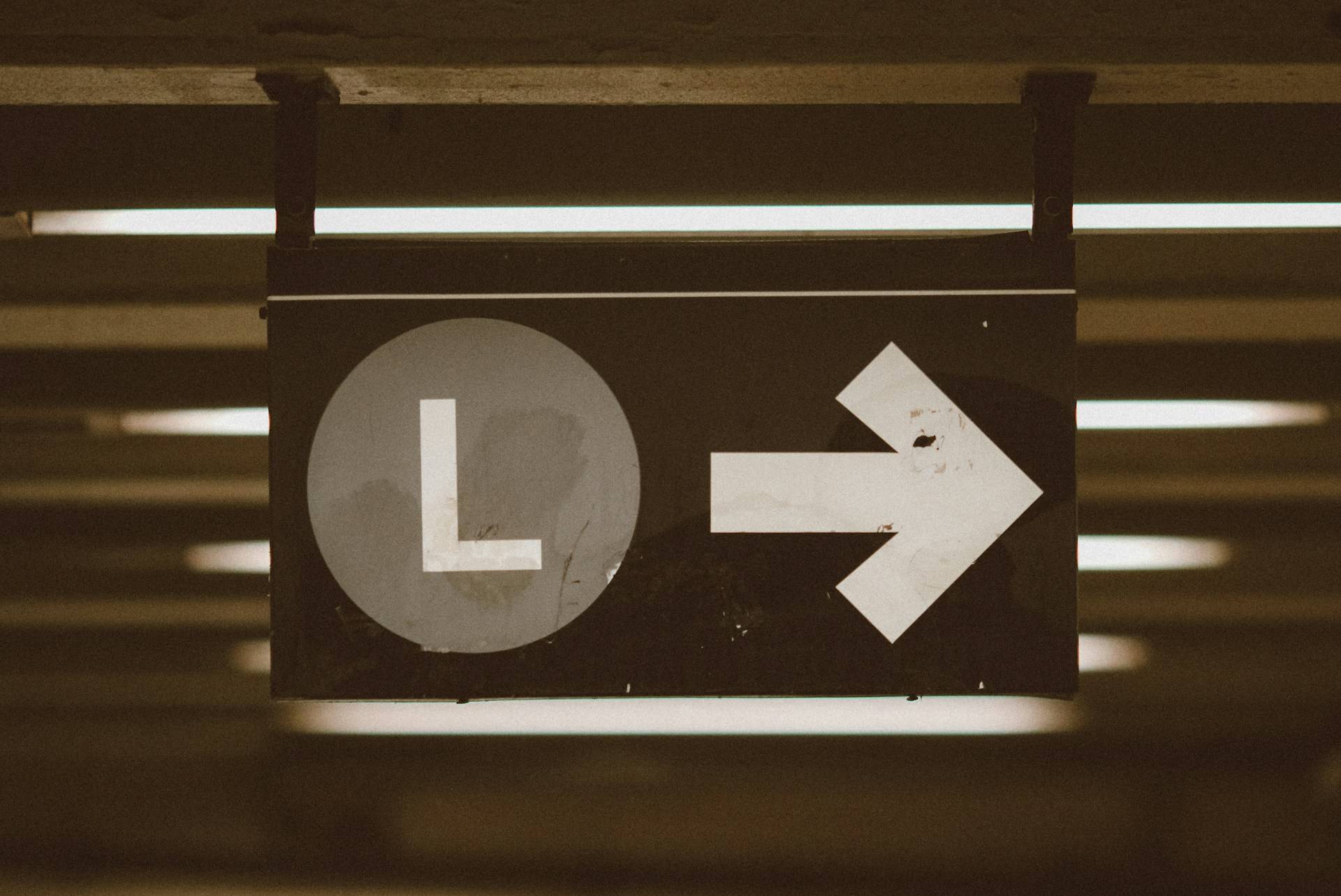 A weathered NYC Metro directional sign hangs from a ceiling in a dimly lit space. The sign features a stark white letter "L" enclosed in a circle on the left side, paired with a bold white arrow pointing right on the right side. Both symbols stand out dramatically against the sign's dark background. The sign shows visible signs of age—scratches and marks that hint at its history of guiding countless wanderers through this space. Fluorescent lights create strips of illumination in the background, casting the utilitarian marker in a nostalgic, slightly melancholic glow that evokes the familiar, sometimes disorienting experience of navigating public spaces.
