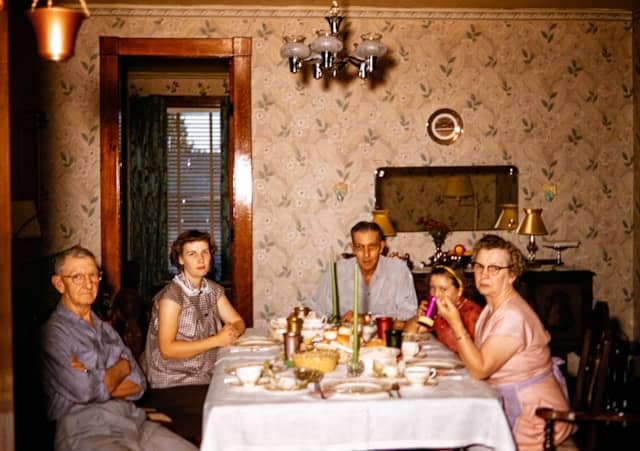 Vintage 35mm photo of a family eating dinner in a dining room. The room is covered in wallpaper and the table suggests this may be a holiday. Five people are gaterhed around in clothes from the 1950s or 1960s and none of them are smiling. 