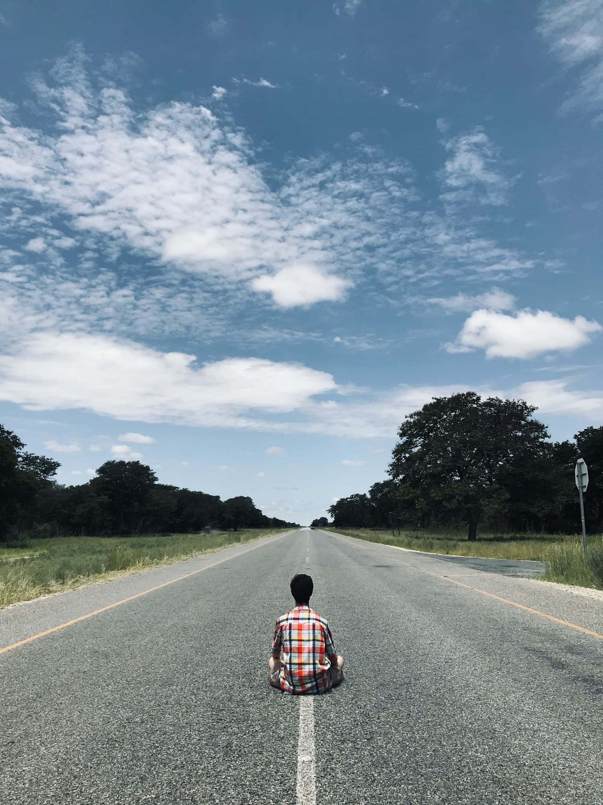 A person in a plaid shirt sits cross-legged in the middle of an empty rural road that stretches toward the horizon, with trees lining both sides and a cloudy blue sky overhead.