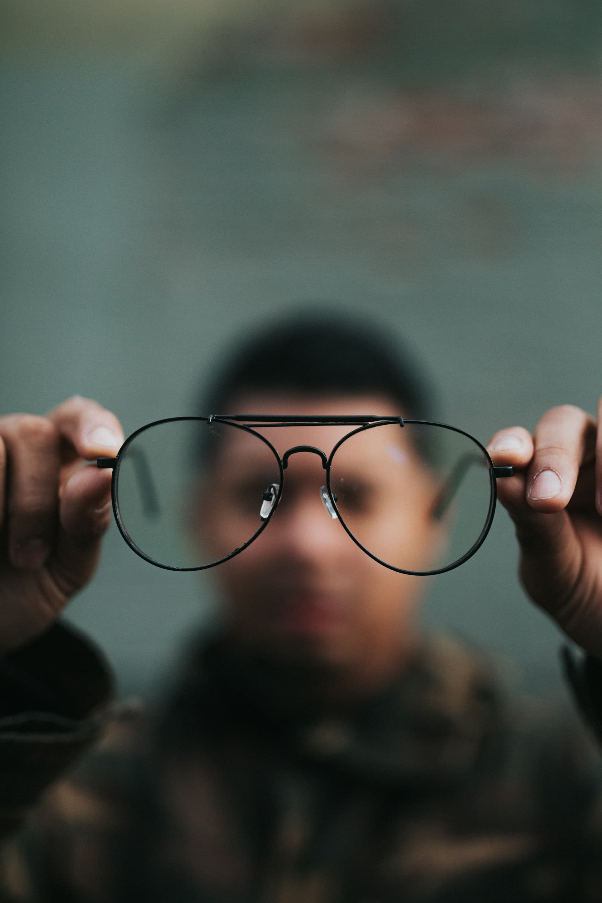 A young man holds a pair of wire-rimmed aviator glasses toward the camera, with the glasses in sharp focus in the foreground while the person's face remains softly blurred in the background.