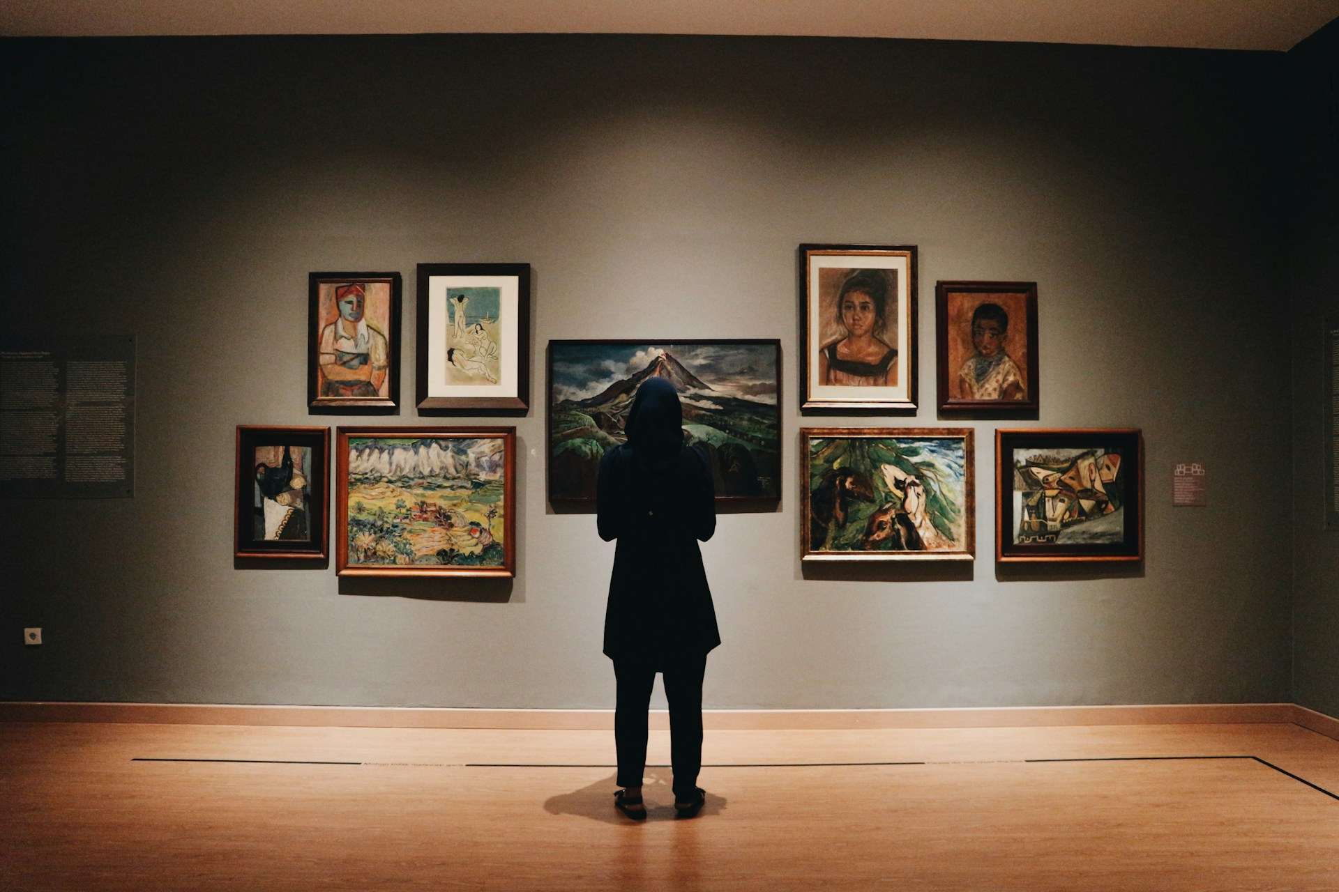 A woman in a black coat and black headscarf stands in front of a wall of paintings inside an art museume in Indonesia. We see her from behind and can only assume she is looking intently at the art. The low lighting has been specially designed to draw atte