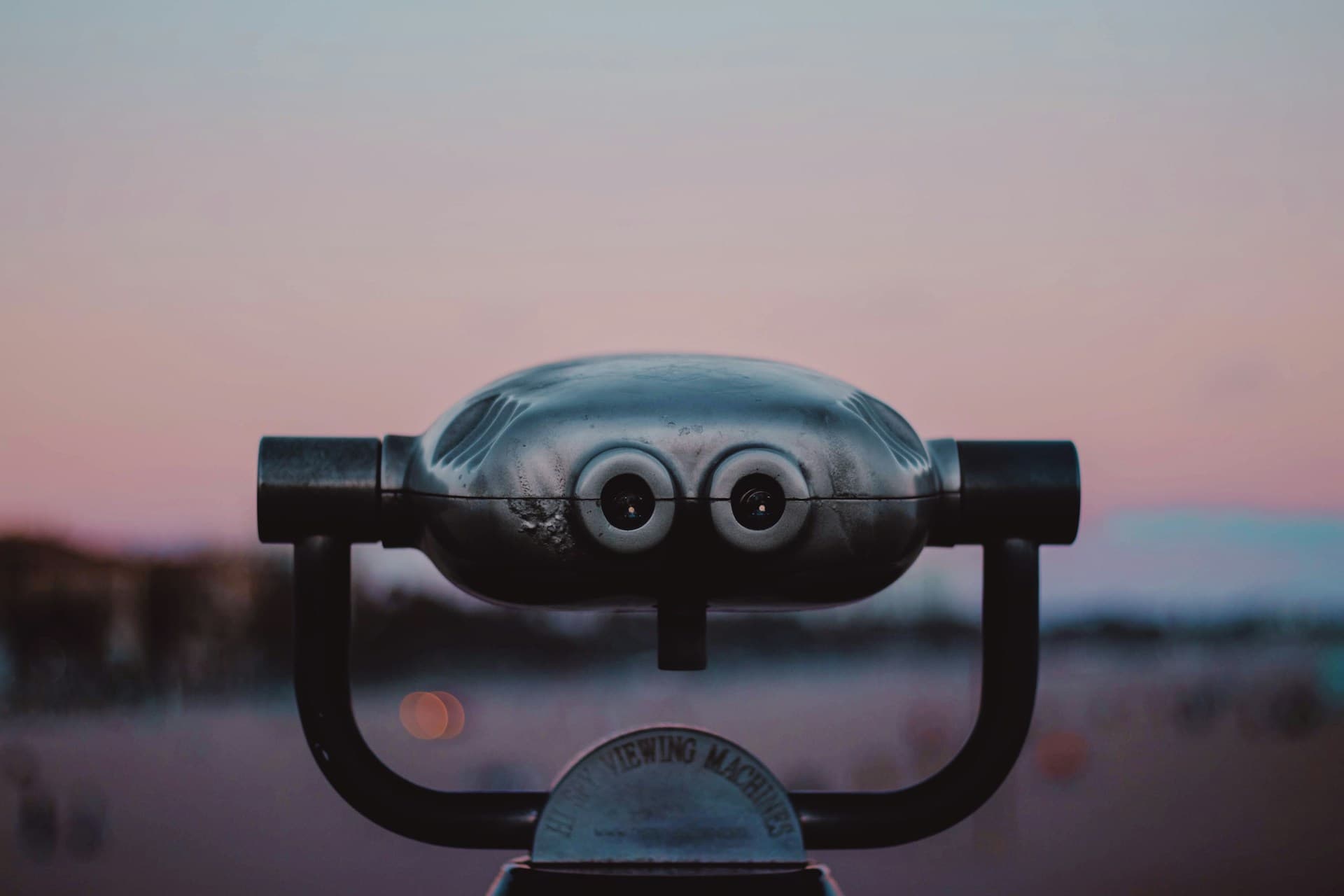 A coin-operated viewing telescope or binoculars mounted on a stand, photographed at dusk or dawn with a soft pink and blue sky in the background. The telescope faces toward a blurred cityscape or landscape in the distance.