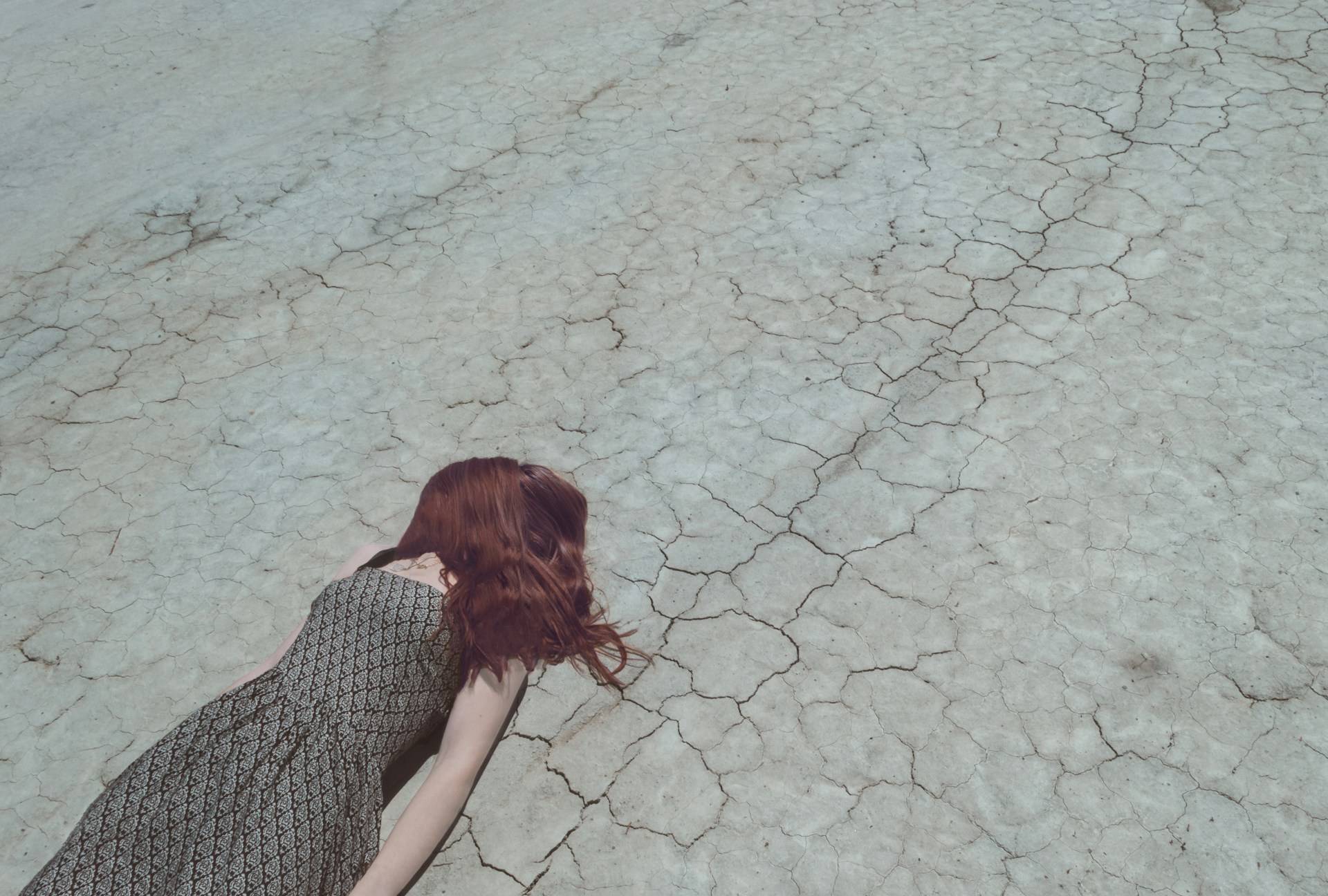 A woman with auburn hair lies face-down on a vast expanse of cracked, pale earth that stretches toward infinity. Her patterned dress contrasts sharply with the parched landscape—a human brushstroke against nature's fractured canvas. The dried mud or salt flat, with its intricate network of fissures resembling a giant's shattered porcelain, dominates the frame while her partial figure anchors the bottom left corner. The composition evokes powerful themes of human vulnerability against environmental desolation, a poetic surrender to forces greater than ourselves. 