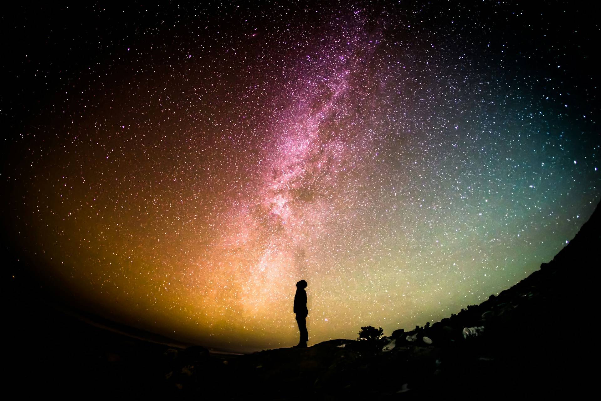A person in black silhouette stands in the middle of a colorful photo of the night sky, where the immensity of the universe overwhelms the human subject. 