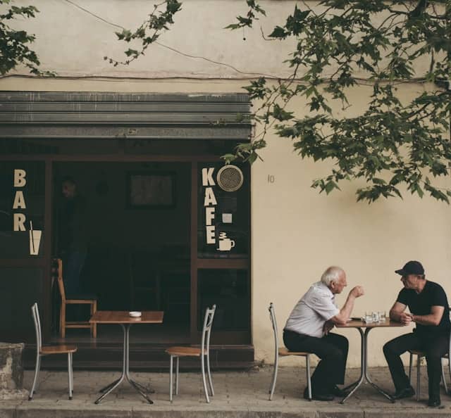 Two old men are having an intense conversation sitting at a table at an outdoor cafe. The photo  was taken in Albania by Juri Gianfrancesco. Photo via unsplash. 