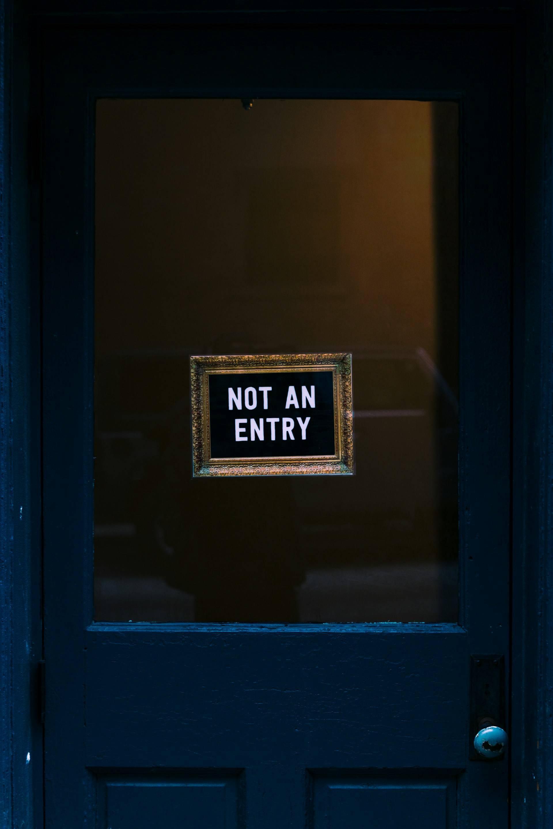 A navy blue door features a rectangular glass panel in its upper portion. Centered within this panel is a framed sign with white text on a black background that reads "NOT AN ENTRY" in capital letters. The sign is displayed in a decorative gold-colored frame with textured edges. The door appears to be part of a darkened interior space, with minimal light illuminating the scene. A metal doorknob is visible in the lower right section of the door. The overall color palette consists primarily of dark blue, black, white, and gold tones. The composition is straightforward—a frontal view of the door with the sign as the focal point.