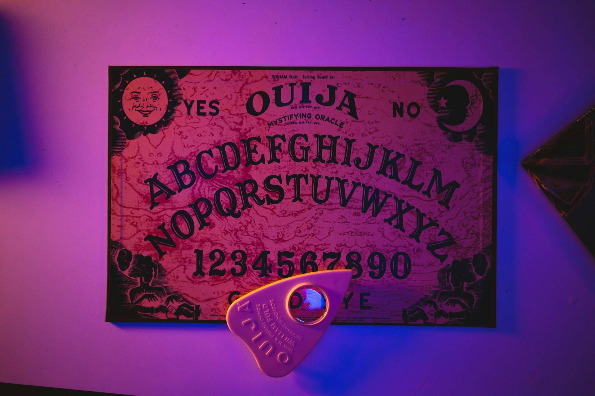 A classic Ouija board with planchette is laid out on a table. The purple-blue lighting of the photo evoke a playful harmlessness. 