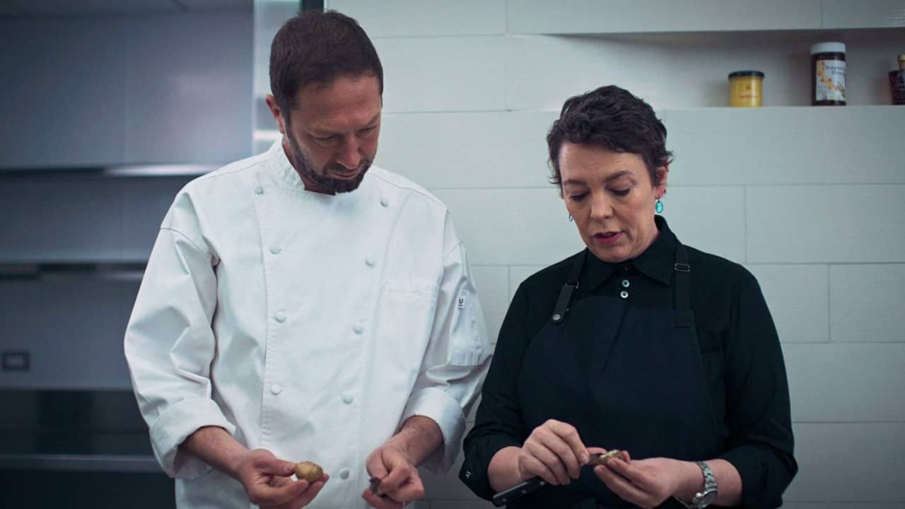A poignant moment captured in a restaurant kitchen as two culinary professionals examine ingredients with intense focus. On the left, Cousin Richie (Eban Moss) in a crisp white double-breasted jacket stands alongside a woman (Olivia Coleman)  in sleek black chef's attire with turquoise earrings. They're both looking down at small food items in their hands, embodying the show's signature attention to detail against a backdrop of clean white tiles. This scene from the acclaimed "Forks" episode illustrates the meticulous care and obsession with perfection that defines high-end hospitality—mirroring the newsletter's exploration of Will Guidara's philosophy of elevating every tiny detail to create transcendent dining experiences.