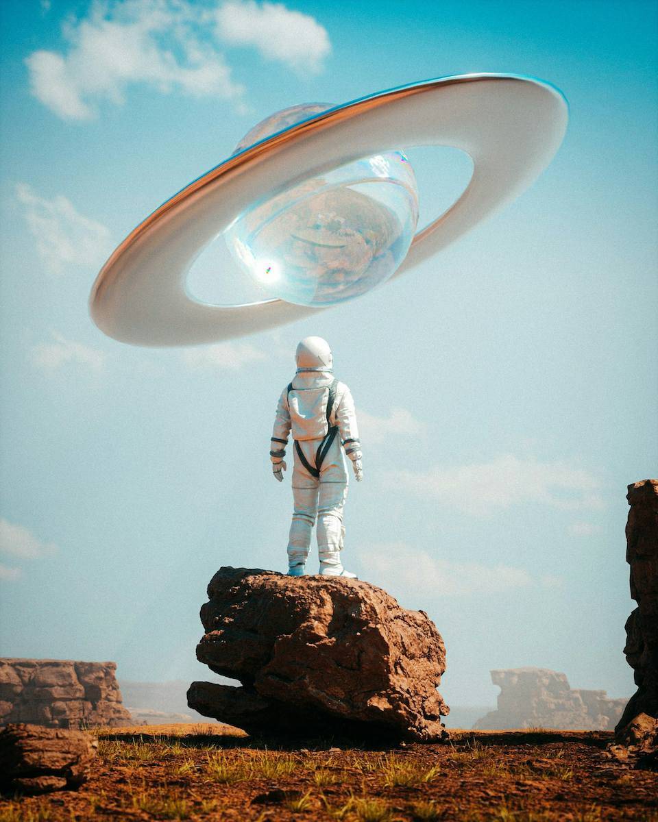 A person wearing a space suit is seen from behind. This person is standing atop a rock on a dry, dusty planet. They look up, and a transluscent Saturn-like planet hangs in the sky. 