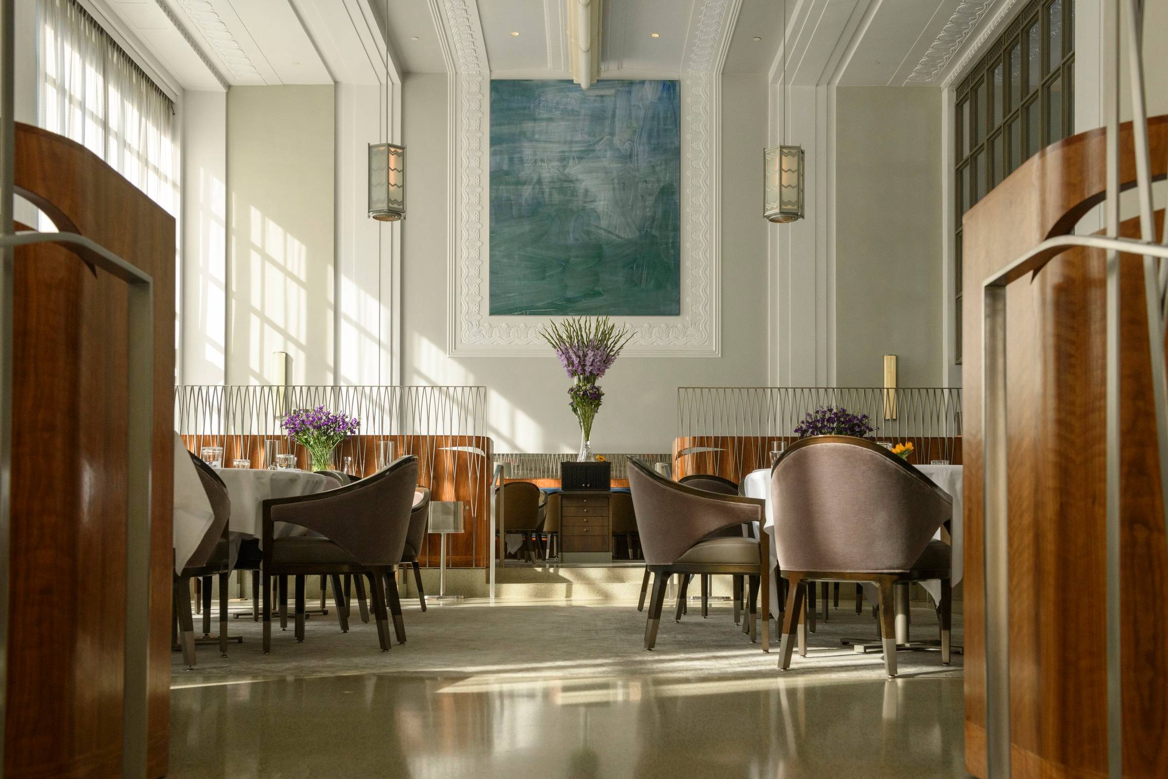 Elegant Modern Restaurant Interior with Artistic Touches  An exquisitely designed restaurant interior viewed through rich wooden doorframes that frame the scene like a living painting. The space marries classical elegance with contemporary flair—crisp white walls adorned with ornate moldings rise to meet a coffered ceiling, while a striking blue-green abstract artwork commands attention on the far wall. Sleek dining tables draped in white linens are surrounded by sophisticated curved-back chairs in a warm taupe hue. Delicate purple flower arrangements punctuate the space, adding vibrant life against the neutral palette. Natural light floods through tall windows on the left, casting dramatic shadows across the polished floor that reflects the entire scene like a mirror. The room employs a thoughtful balance of textures—wood, fabric, metal, and glass—creating an atmosphere that's both luxurious and inviting, where culinary artistry likely matches the visual sophistication of the space itself.