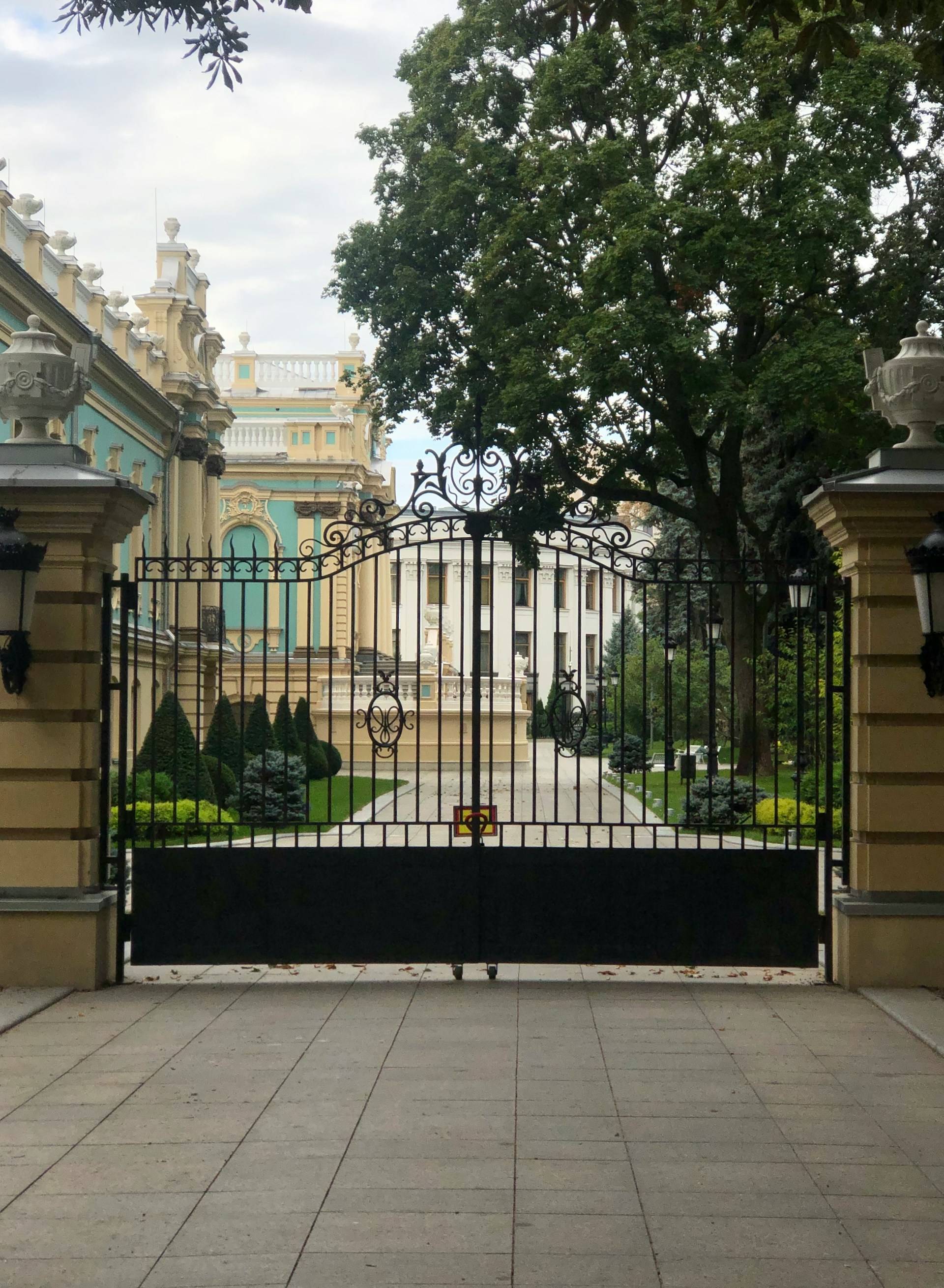 An ornate wrought iron gate stands closed at the entrance to a grand historic building. The gate features decorative scrollwork and is mounted between two substantial stone pillars with ornamental urns at the top. Beyond the gate is a paved driveway leading to an elegant palace or mansion with turquoise and cream-colored façade elements. The building displays classical architectural features including balustrades and decorative moldings. The grounds are meticulously landscaped with shaped conical evergreen shrubs and manicured lawns on either side of the path. Mature trees frame the scene, with their dense green foliage extending across the upper portion of the image. The foreground shows a stone or concrete paved area leading up to the gate. The sky is lightly clouded, creating a soft, diffused light over the entire scene.