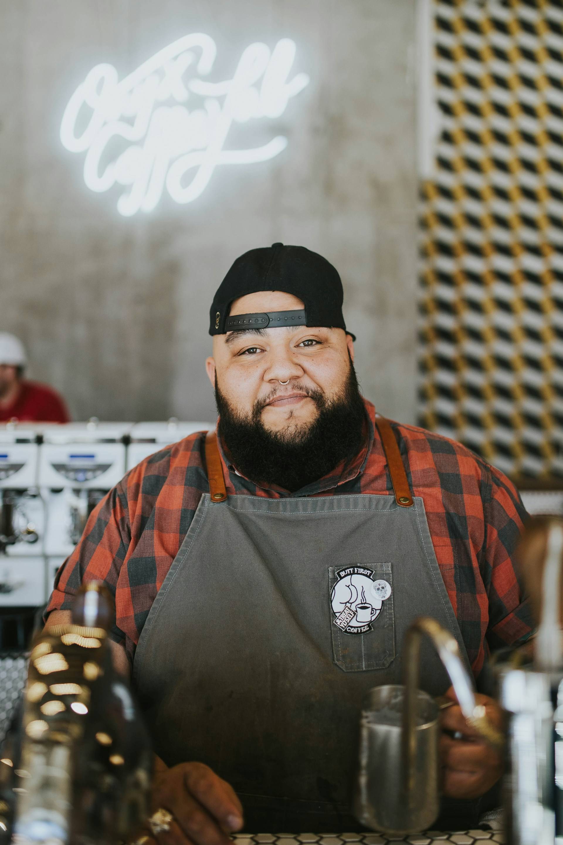 A warm, inviting coffee shop scene featuring a friendly barista standing behind the counter with a genuine smile. The barista wears a backwards black cap, orange and black plaid flannel shirt, and a gray apron with a coffee-themed logo patch.