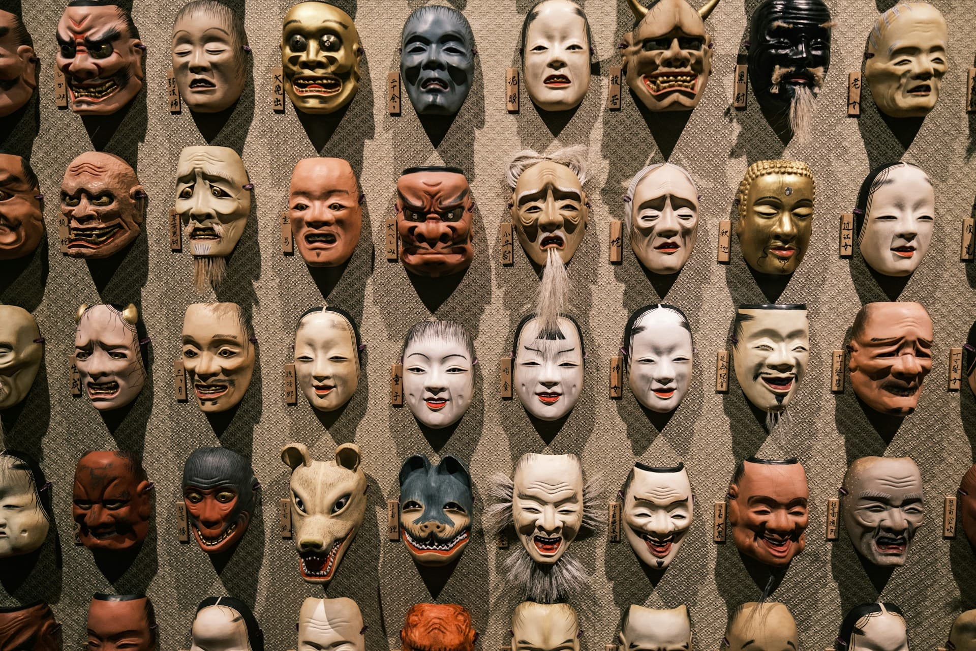 A collection of traditional theatrical masks displayed on a woven mat or wall, showing various expressions and characters. The masks appear to be Asian in origin, possibly Japanese Noh or similar traditional theater masks, with different facial expressions ranging from serene to dramatic.