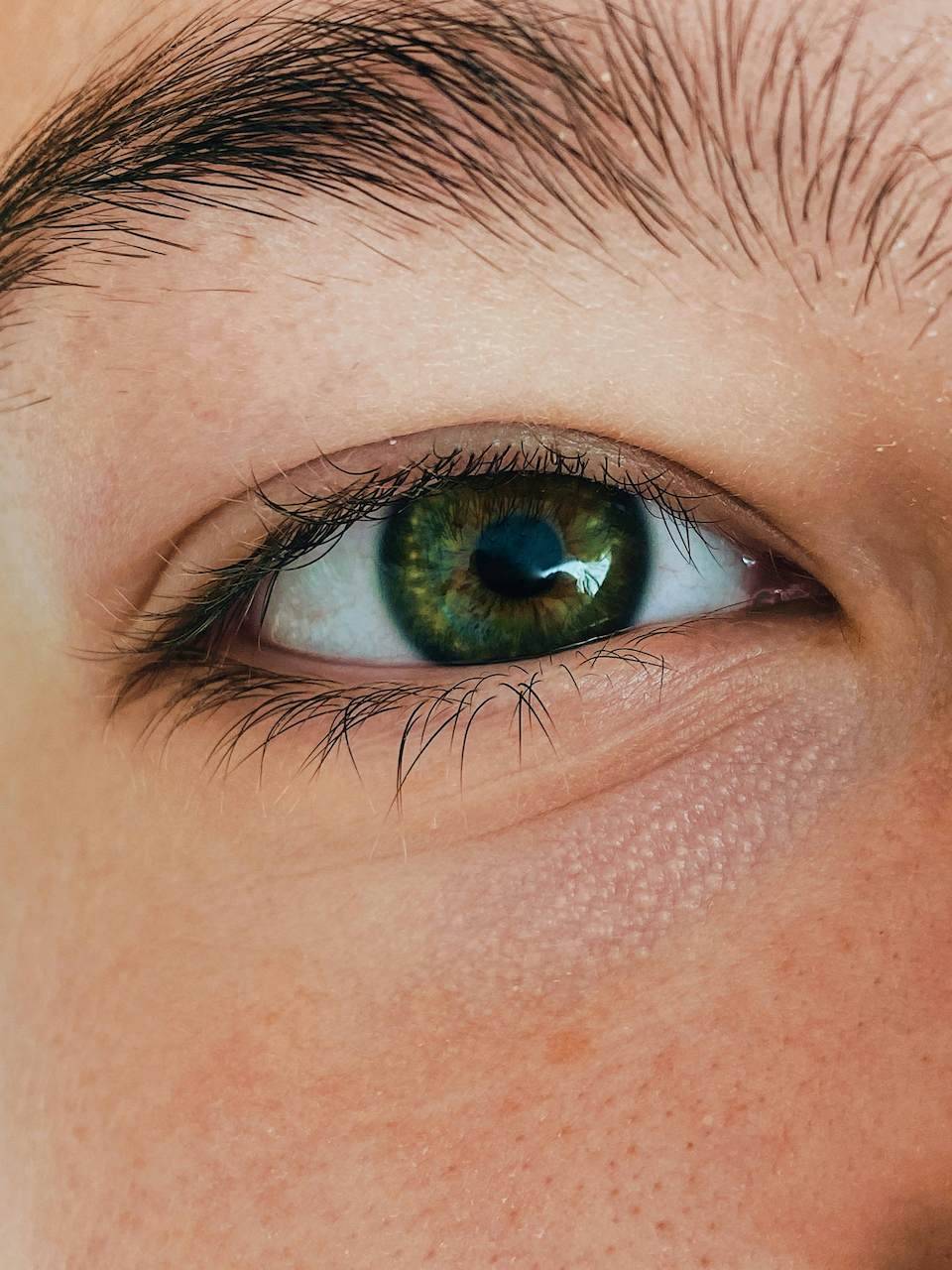 Close up of a human eye, dark green in color with patches of dark brown. The eye is surrounded by dark eyelashes and a dark eyebrow. The close-up image allows us to see pores in the skin just under the eye as well as freckles and red spots. Photo by Severin Demchuk via Unsplash