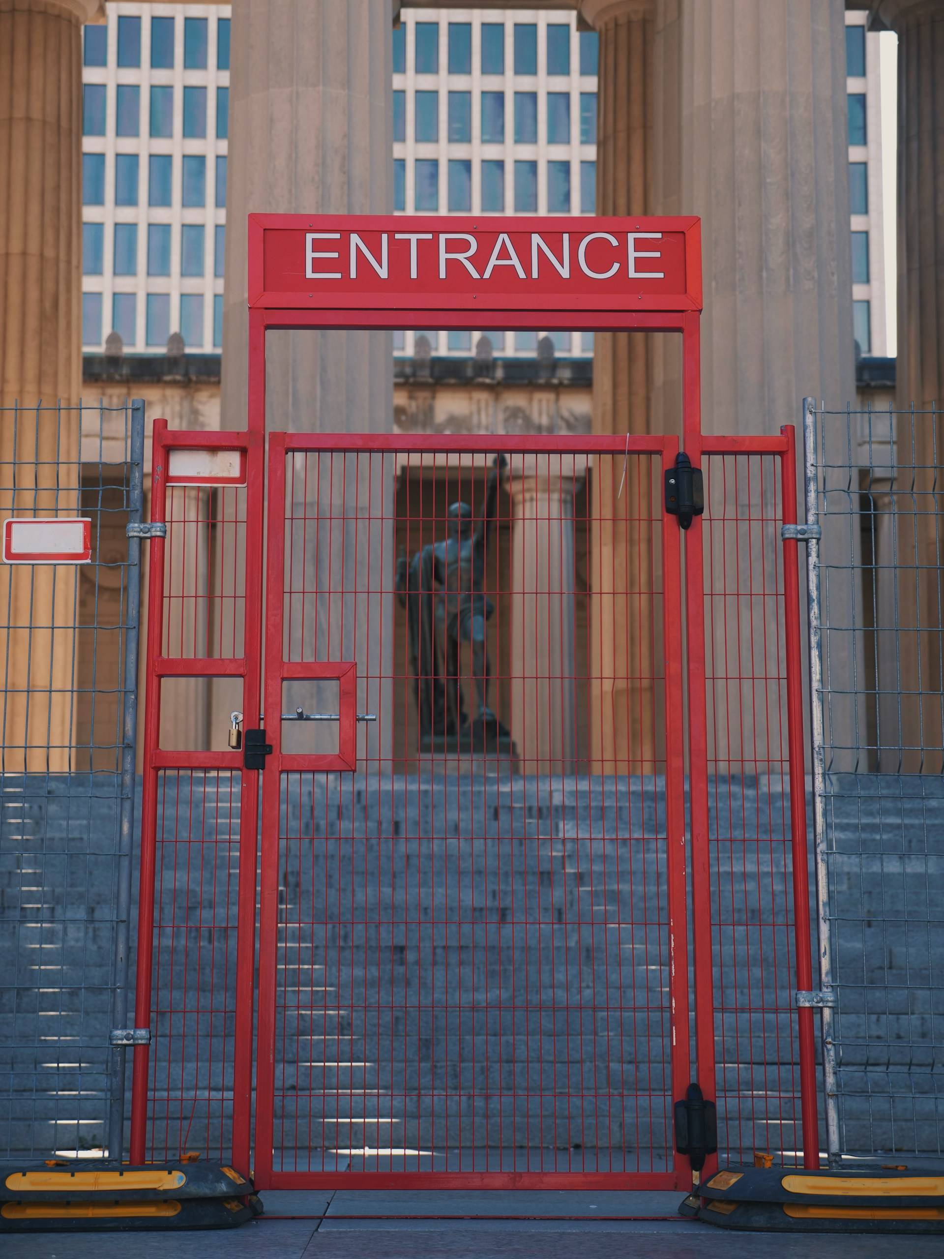 A bright red metal gate marks the entrance to a classical building with stone columns. The gate features a prominent header bar with the word "ENTRANCE" displayed in white lettering. The gate consists of vertical bars and has a small door within its structure with visible locks and hinges. Temporary mesh fencing connects to either side of the entrance structure. The gate sits at the top of several stone steps. Behind the gate, partially visible, is what appears to be a statue inside the building entrance. The background shows large stone columns typical of neoclassical architecture, with a modern office building visible through the spaces between the columns. Yellow safety bumpers are placed at the base of the gate on the ground.