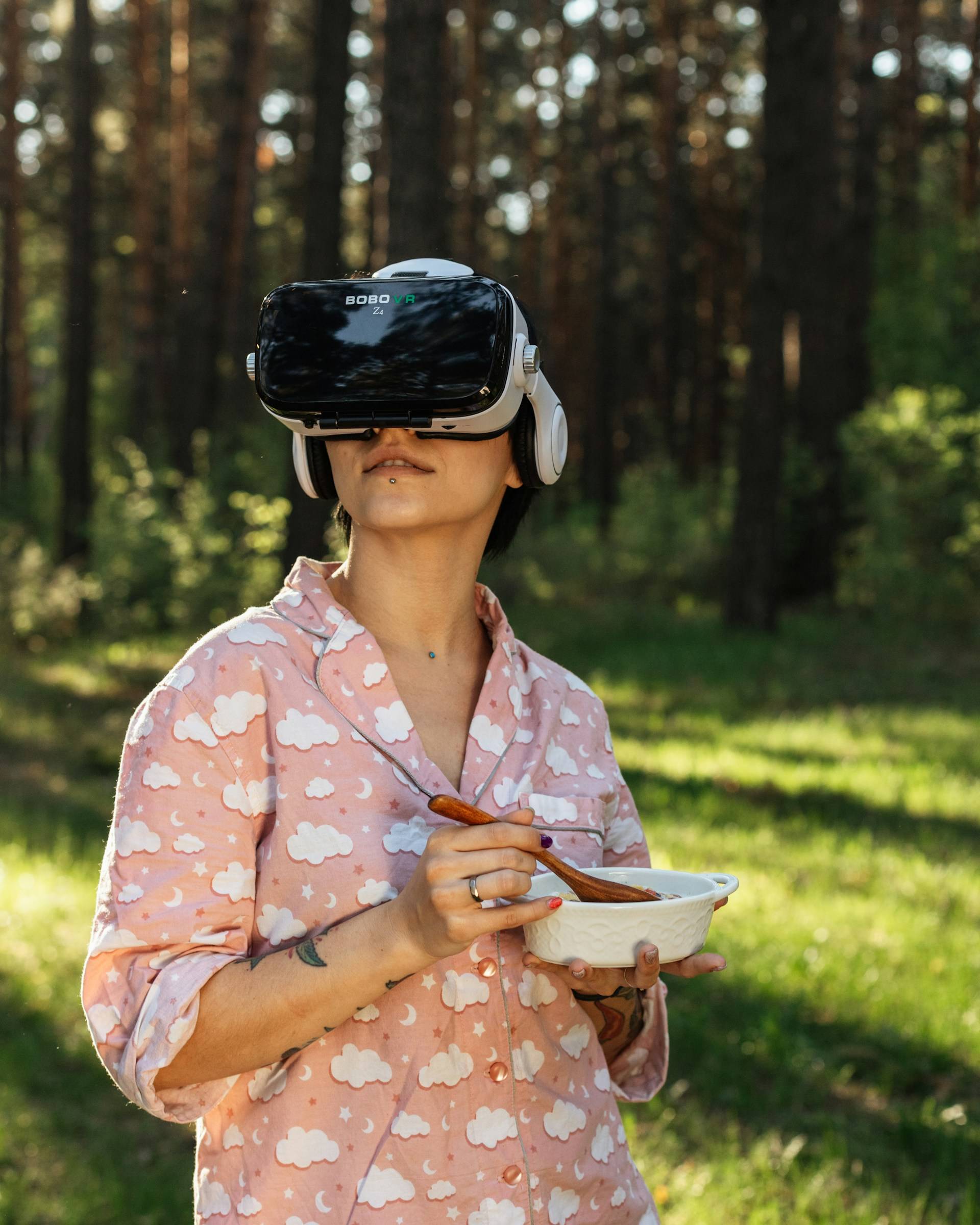 A young woman wearing pink pajamas with fluffy white clouds is standing outside in the morning light. She's wearing a full VR headset while eating cereal out of a bowl. The woman sports a pierced chin and a simple wedding band. 