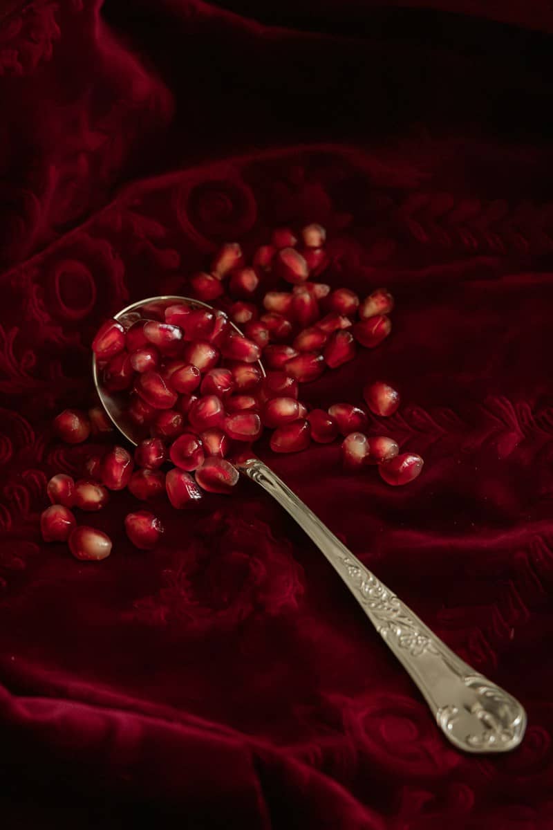 A silver spoon overflowing with pomegranate seeds on red fabric.