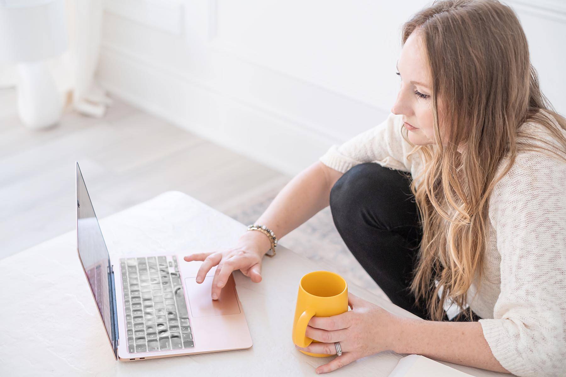 Rebecca Joy of joybeforework.com is wearing a cream top and black pants, typing on a laptop, hot beverage in hand