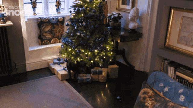 gif of a black dog at the bottom of a christmas tree decorate with lights and blue baubles. He ferrets around in the bottom of the tree, topples it over and runs off before it lands on him.