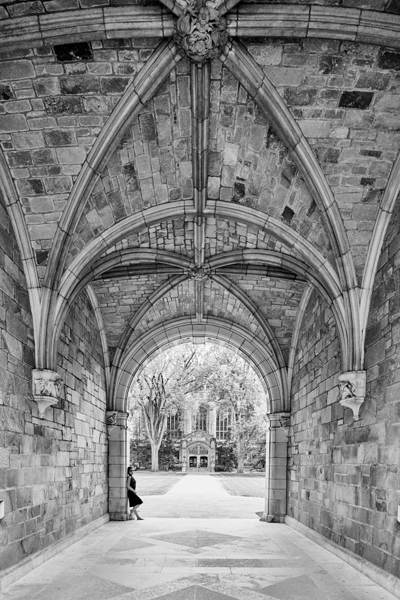 A black-and-white image of a beautiful arched stone walkway with intricate Gothic architecture, featuring a woman in a black dress leaning against one of the pillars. Beyond the archway, a courtyard opens up to a grand building framed by large trees.