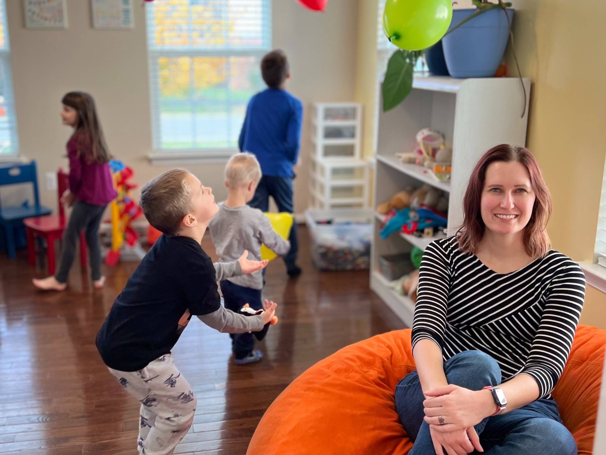 Autumn smiles while seated in a cozy, sunlit room as her four children play energetically around her—a real-life glimpse into the joy and chaos of parenting.