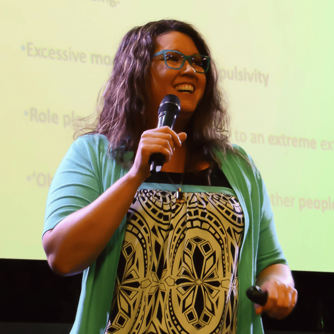 An image of Christina Keeble, a fair skinned woman with brown, curly hair, holding a microphone, wearing teal glasses, & smiling as she stands in front of a screen with her PowerPoint projected onto it during a presentation she gave.