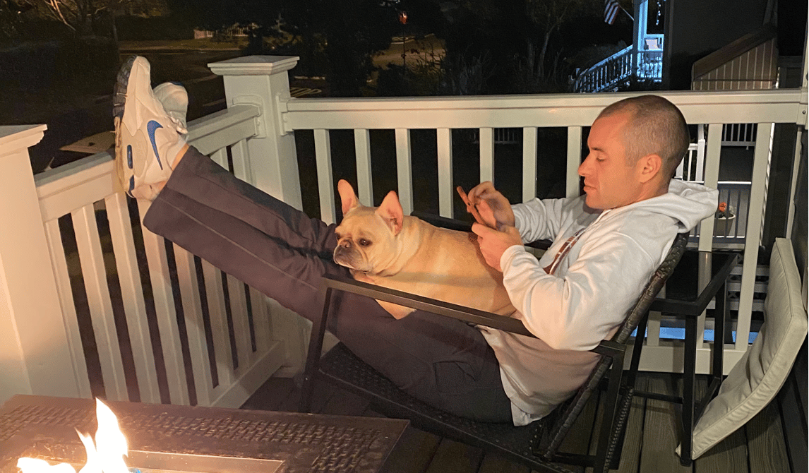 Jeff Proctor, co-founder of DollarSprout, relaxing on a porch with his dog while scrolling on his phone by a fire pit at night.