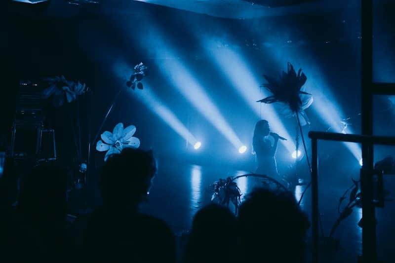 A person singing into a microphone on a stage with large artificial flowers